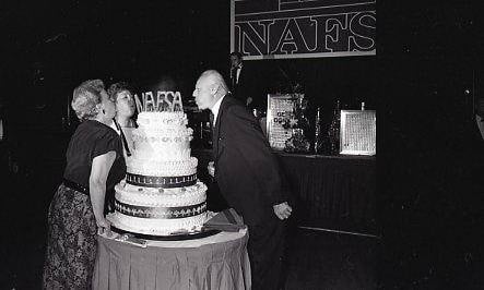 Black and white image of two people blowing candles out on a large cake